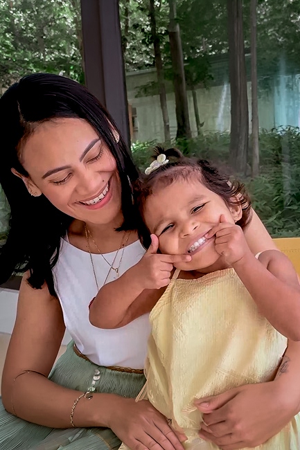 Carolina del Valle smiles tenderly as she hugs her two-year-old daughter, Aitana Banda. The little girl looks at the camera with joy, playfully stretching her cheeks and showing her teeth. They are in a brightly lit space with trees in the background.