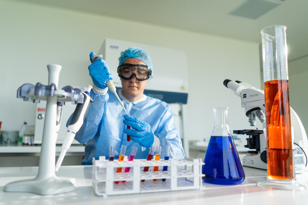 Un especialista realiza pruebas con una pipeta de precisión en el laboratorio del Hospital Serena del Mar, reconocido por Great Place to Work Colombia. El profesional viste equipo de bioprotección completo y está rodeado de instrumental científico como tubos de ensayo y matraces con líquidos coloridos, simbolizando la excelencia y tecnología de la institución.