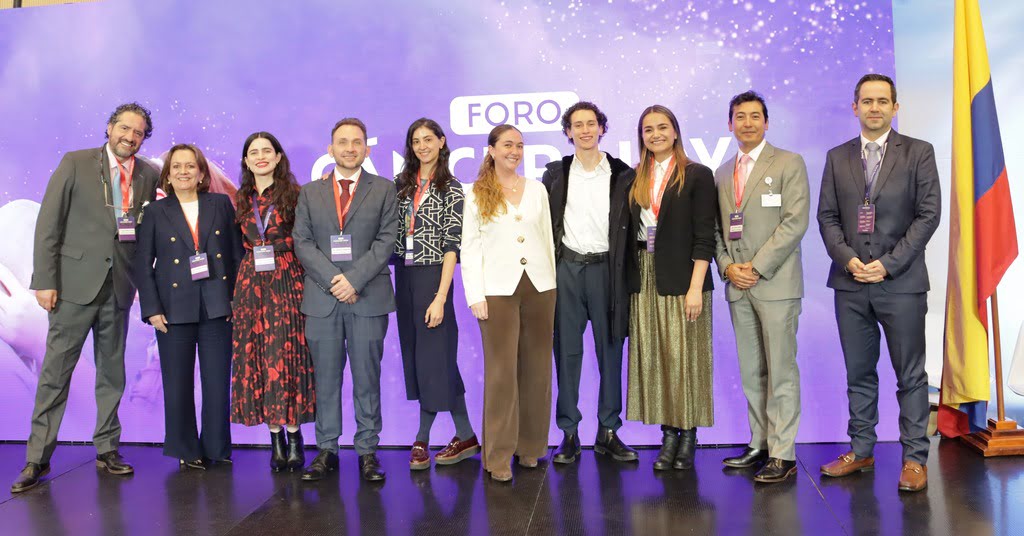 Diez profesionales (cinco mujeres y cinco hombres) sonríen en el evento 'Cáncer hoy: retos y futuro'. Visten ropa formal con credenciales ante un fondo violeta con la palabra 'FORO'. A la derecha se ve la bandera de Colombia. El grupo representa a los especialistas líderes del encuentro.
