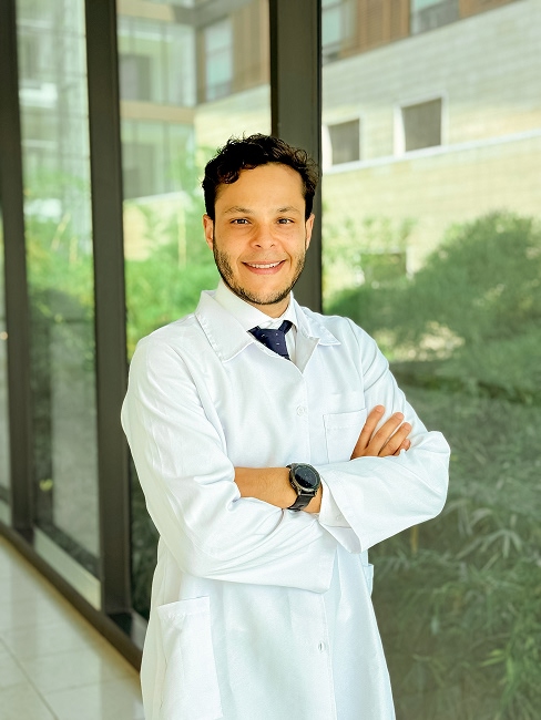 Retrato del Dr. Andrés Felipe Porto Mercado, médico del Hospital Serena del Mar, vistiendo bata blanca, camisa y corbata. El Dr. Porto, egresado de la Universidad del Rosario y especialista de la FUCS, posa sonriente con los brazos cruzados en un pasillo moderno y luminoso del hospital.