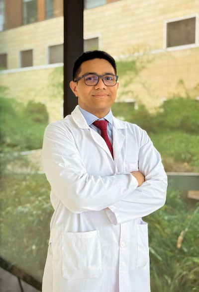 Retrato profesional del Dr. Pedro Caraballo Gómez, especialista en Hematología Oncológica. El doctor aparece en un plano medio, vistiendo una bata médica blanca sobre una camisa azul claro y una corbata roja. Se encuentra con los brazos cruzados y una expresión amable y segura, usando anteojos de montura oscura. El fondo es un entorno exterior luminoso con un edificio de arquitectura moderna y vegetación difuminada.