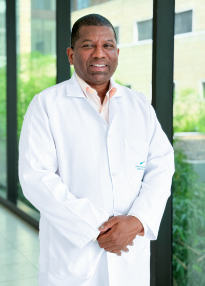 Retrato profesional del Dr. Julio de la Rosa Adonilso, especialista en Psiquiatría. El doctor aparece de pie, vistiendo una bata médica blanca impecable sobre una camisa de cuello claro, con las manos cruzadas al frente y una expresión serena y confiable. La fotografía está tomada en un entorno clínico moderno y luminoso, con ventanales de fondo que muestran follaje verde difuminado.