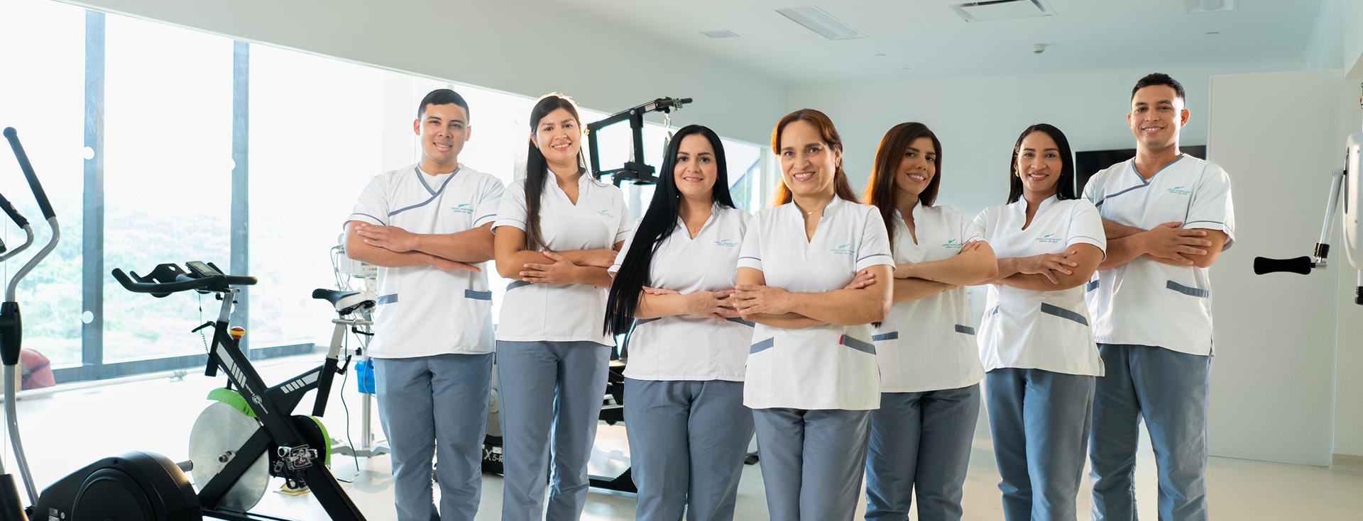 Siete profesionales de la salud del Hospital Serena del Mar, cuatro mujeres y tres hombres, posan con confianza y una sonrisa amable en un moderno gimnasio de rehabilitación. El equipo viste uniformes institucionales compuestos por camisas blancas con detalles grises y pantalones azul claro. La imagen refleja un ambiente de trabajo colaborativo, profesional y humano.