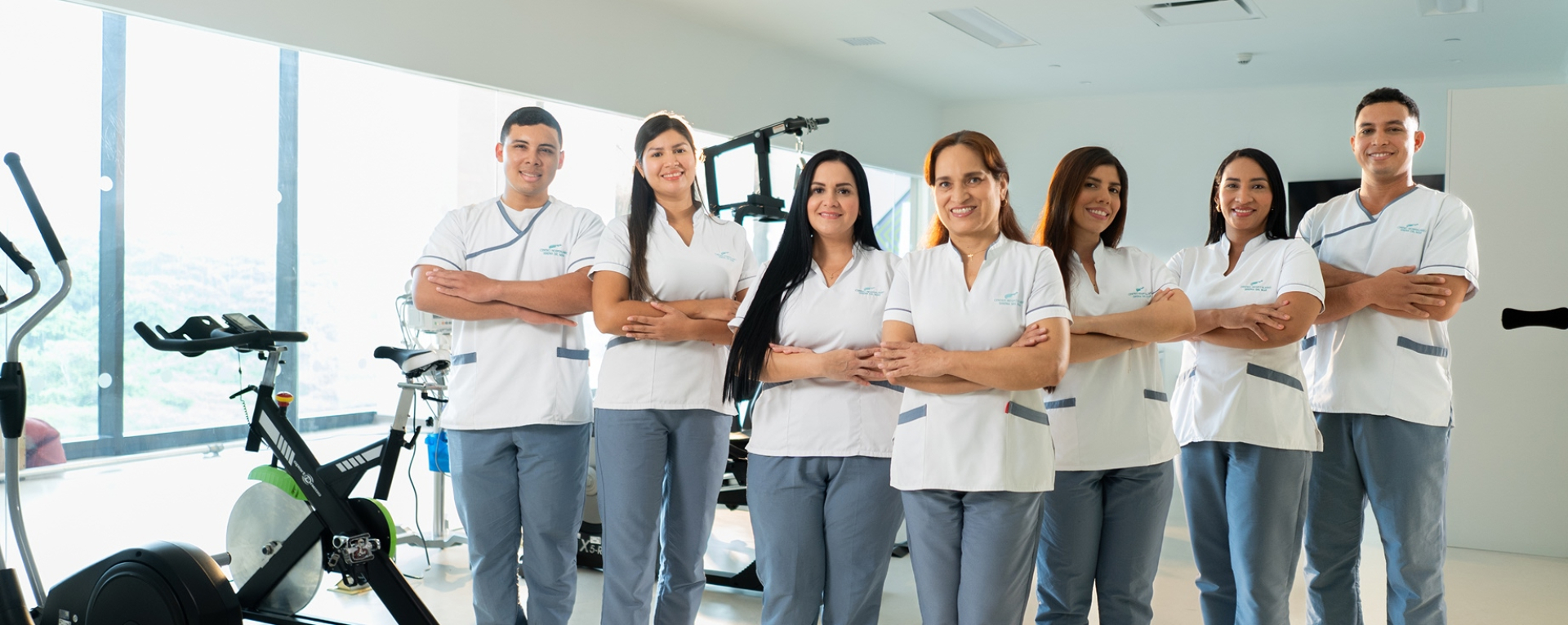 Siete profesionales de la salud del Hospital Serena del Mar, cuatro mujeres y tres hombres, posan con confianza y una sonrisa amable en un moderno gimnasio de rehabilitación. El equipo viste uniformes institucionales compuestos por camisas blancas con detalles grises y pantalones azul claro. La imagen refleja un ambiente de trabajo colaborativo, profesional y humano.