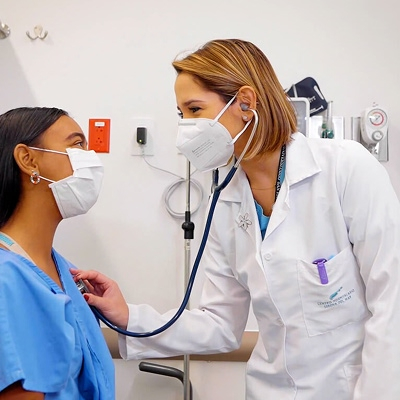 Una doctora del Hospital Serena del Mar sonríe cálidamente mientras utiliza su estetoscopio para atender a una paciente. Ambas usan mascarillas en un consultorio moderno, reflejando un ambiente de cuidado profesional y confianza.