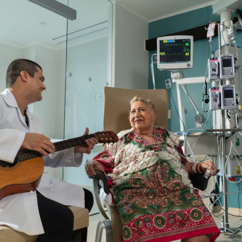 Un profesional de la salud con bata blanca toca la guitarra para una paciente adulta mayor en una habitación del Hospital Serena del Mar, institución certificada como UCI Eficiente por Epimed. La paciente, vestida con una colorida manta tradicional, sonríe con alegría mientras lo escucha. Al fondo, se observa equipo médico de alta tecnología, incluyendo monitores de signos vitales y bombas de infusión, resaltando la combinación de humanización del servicio y eficiencia clínica.