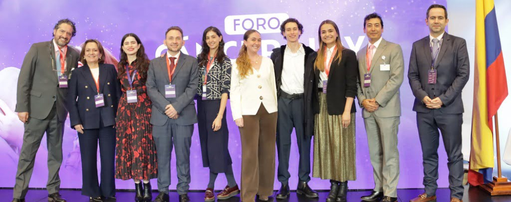 Diez profesionales (cinco mujeres y cinco hombres) sonríen en el evento 'Cáncer hoy: retos y futuro'. Visten ropa formal con credenciales ante un fondo violeta con la palabra 'FORO'. A la derecha se ve la bandera de Colombia. El grupo representa a los especialistas líderes del encuentro.
