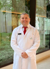 Retrato profesional del Dr. Luis Gabriel Salgado Montiel, especialista en Medicina Interna. El doctor posa de pie con una bata blanca de laboratorio sobre una camisa blanca y una corbata roja con detalles blancos. Mantiene una expresión sonriente y amable, con las manos entrelazadas al frente. El fondo muestra un ventanal con plantas verdes, creando un ambiente clínico iluminado y tranquilo.