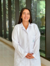 Retrato de medio cuerpo de la Dra. María Mónica Valencia Murillo, especialista en Geriatría del Hospital Serena del Mar. Viste una bata blanca de laboratorio con el logotipo del hospital bordado en el bolsillo superior. La doctora, de cabello largo oscuro y expresión amable, posa de pie en un pasillo moderno y luminoso con ventanales de fondo.