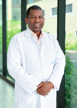 Retrato profesional del Dr. Julio de la Rosa Adonilso, especialista en Psiquiatría. El doctor aparece de pie, vistiendo una bata médica blanca impecable sobre una camisa de cuello claro, con las manos cruzadas al frente y una expresión serena y confiable. La fotografía está tomada en un entorno clínico moderno y luminoso, con ventanales de fondo que muestran follaje verde difuminado.