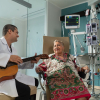 Un profesional de la salud con bata blanca toca la guitarra para una paciente adulta mayor en una habitación del Hospital Serena del Mar, institución certificada como UCI Eficiente por Epimed. La paciente, vestida con una colorida manta tradicional, sonríe con alegría mientras lo escucha. Al fondo, se observa equipo médico de alta tecnología, incluyendo monitores de signos vitales y bombas de infusión, resaltando la combinación de humanización del servicio y eficiencia clínica.