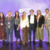 Diez profesionales (cinco mujeres y cinco hombres) sonríen en el evento 'Cáncer hoy: retos y futuro'. Visten ropa formal con credenciales ante un fondo violeta con la palabra 'FORO'. A la derecha se ve la bandera de Colombia. El grupo representa a los especialistas líderes del encuentro.