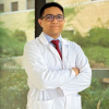 Retrato profesional del Dr. Pedro Caraballo Gómez, especialista en Hematología Oncológica. El doctor aparece en un plano medio, vistiendo una bata médica blanca sobre una camisa azul claro y una corbata roja. Se encuentra con los brazos cruzados y una expresión amable y segura, usando anteojos de montura oscura. El fondo es un entorno exterior luminoso con un edificio de arquitectura moderna y vegetación difuminada.