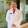 Retrato profesional del Dr. Luis Gabriel Salgado Montiel, especialista en Medicina Interna. El doctor posa de pie con una bata blanca de laboratorio sobre una camisa blanca y una corbata roja con detalles blancos. Mantiene una expresión sonriente y amable, con las manos entrelazadas al frente. El fondo muestra un ventanal con plantas verdes, creando un ambiente clínico iluminado y tranquilo.