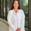 Retrato de medio cuerpo de la Dra. María Mónica Valencia Murillo, especialista en Geriatría del Hospital Serena del Mar. Viste una bata blanca de laboratorio con el logotipo del hospital bordado en el bolsillo superior. La doctora, de cabello largo oscuro y expresión amable, posa de pie en un pasillo moderno y luminoso con ventanales de fondo.