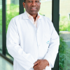 Retrato profesional del Dr. Julio de la Rosa Adonilso, especialista en Psiquiatría. El doctor aparece de pie, vistiendo una bata médica blanca impecable sobre una camisa de cuello claro, con las manos cruzadas al frente y una expresión serena y confiable. La fotografía está tomada en un entorno clínico moderno y luminoso, con ventanales de fondo que muestran follaje verde difuminado.